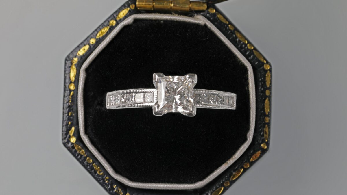 18K white gold princess cut diamond ring displayed in a ring box featuring a brilliant princess cut center diamond accented by channel set diamonds in a sleek modern setting.