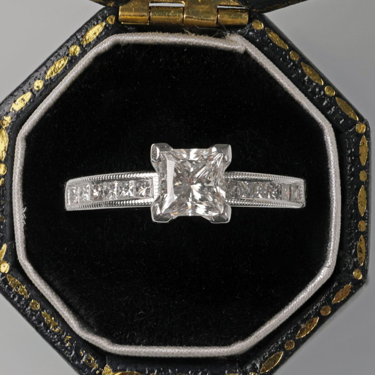 18K white gold princess cut diamond ring displayed in a ring box featuring a brilliant princess cut center diamond accented by channel set diamonds in a sleek modern setting.