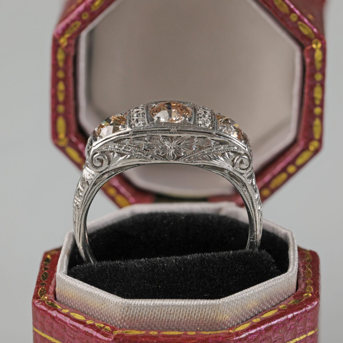 Side view of a Platinum Art Deco style ring displayed in a ring box featuring three light brown old cut diamonds in an ornate vintage inspired setting with additional accent diamonds.