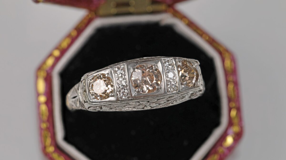 Platinum Art Deco style ring displayed in a ring box featuring three light brown old cut diamonds in an ornate vintage inspired setting with additional accent diamonds.