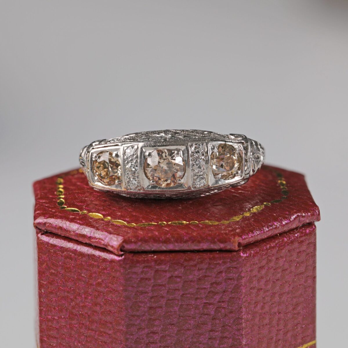 Platinum Art Deco style ring displayed sitting on a red ring box featuring three light brown old cut diamonds in an ornate vintage inspired setting with additional accent diamonds.