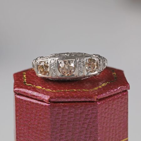 Platinum Art Deco style ring displayed sitting on a red ring box featuring three light brown old cut diamonds in an ornate vintage inspired setting with additional accent diamonds.