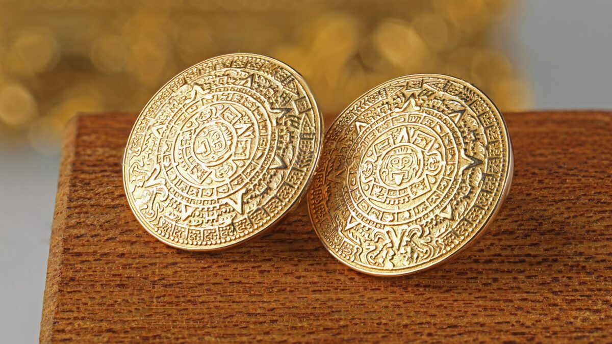 Pair of retro 18K yellow gold Aztec disc earrings sitting on a wooden box featuring bold geometric circular designs inspired by ancient motifs and sculptural styling.