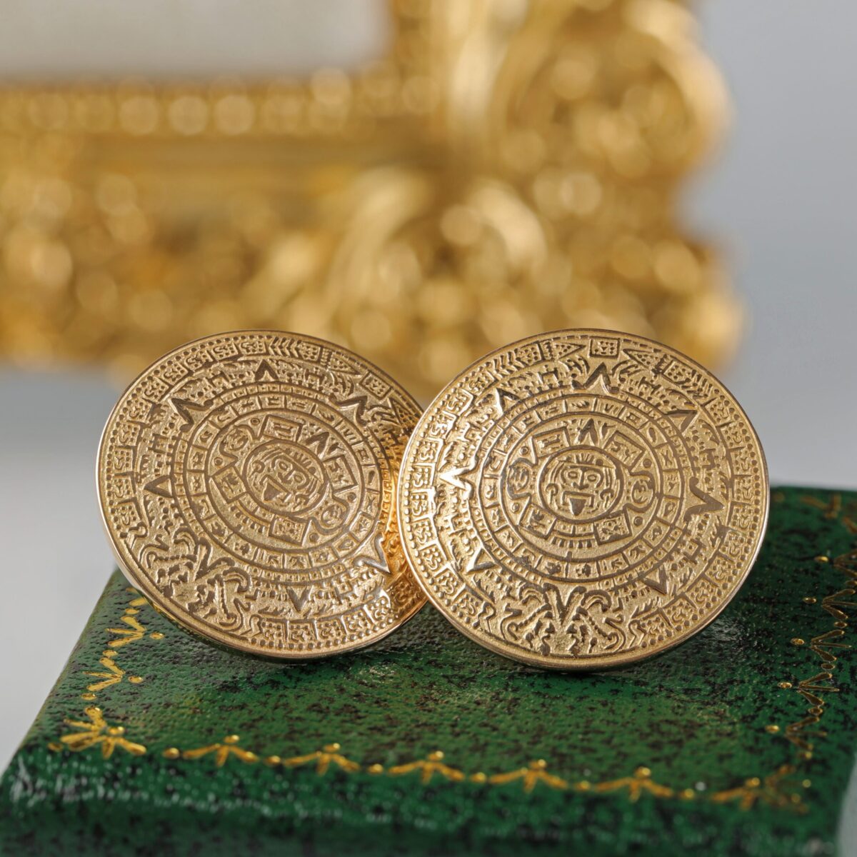 Pair of retro 18K yellow gold Aztec disc earrings sitting on a green box featuring bold geometric circular designs inspired by ancient motifs and sculptural styling.
