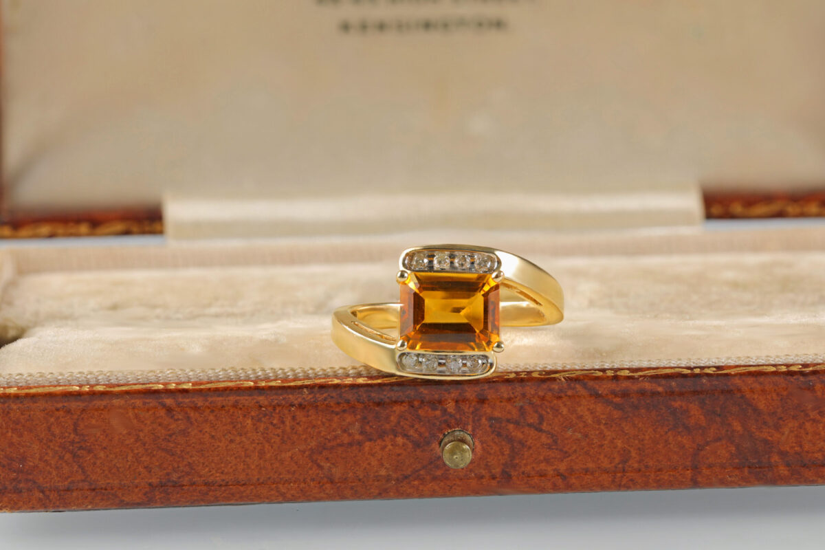 Citrine bypass ring displayed in a jewelry box featuring an emerald cut orangy-yellow citrine accented by round brilliant diamonds in 14K yellow gold.