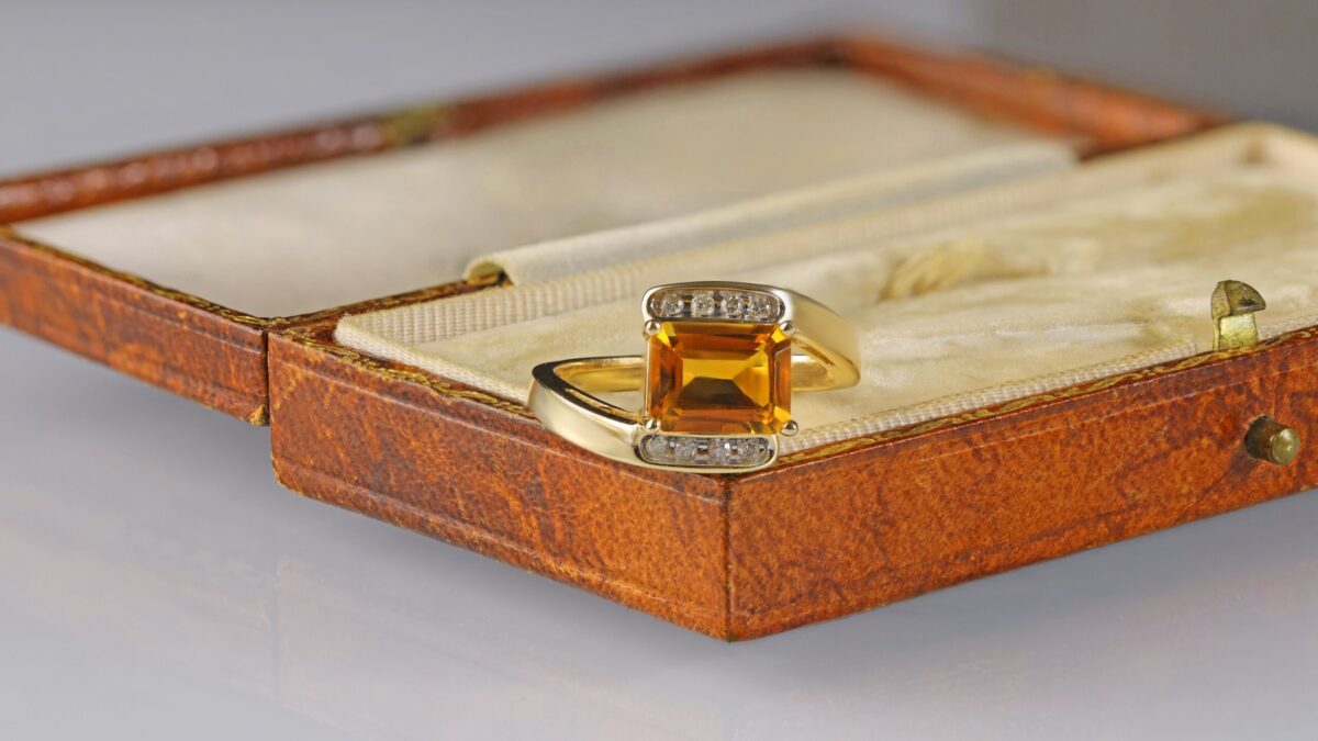 Citrine bypass ring displayed in a jewelry box featuring an emerald cut orangy-yellow citrine accented by round brilliant diamonds in 14K yellow gold.