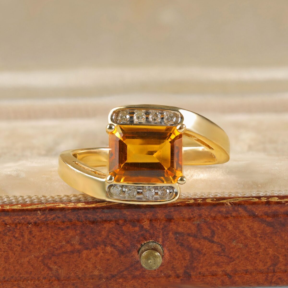 Citrine bypass ring displayed in a jewelry box featuring an emerald cut orangy-yellow citrine accented by round brilliant diamonds in 14K yellow gold.