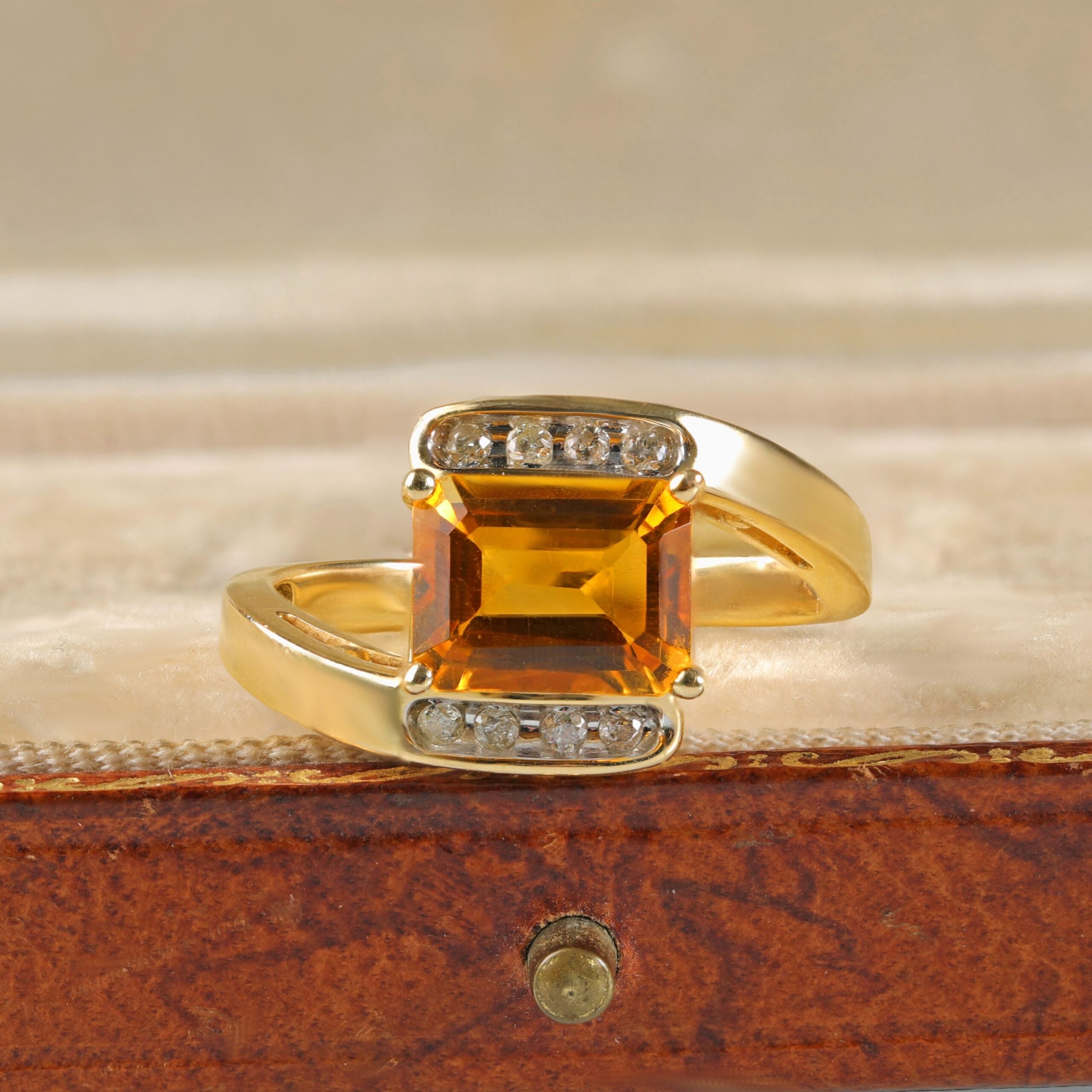 Citrine bypass ring displayed in a jewelry box featuring an emerald cut orangy-yellow citrine accented by round brilliant diamonds in 14K yellow gold.
